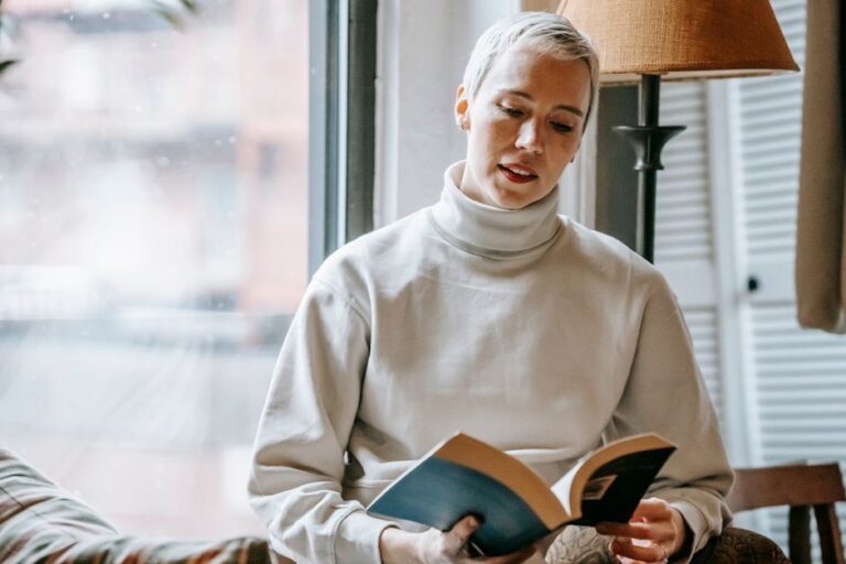 Intelligent content female reading volume of fascinating book near window of lounge in daytime